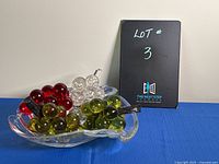 Lucite grapes in red, clear, and green colors on swirl pattern glass bowl photographed against a neutral background and blue cloth surface.