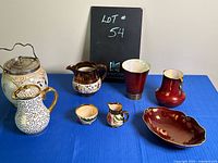 Front view showing all seven assorted ceramic and pottery items on a blue cloth background with a small blackboard showing lot number