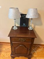 Front view of the walnut nightstand with one drawer and cabinet door, showing two glass stem lamps with white textured shades on top.