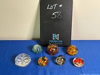 Front view of the seven glass paperweights showing various swirl and floral designs, placed on a blue cloth with black binder behind.