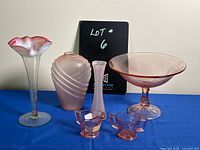 Wide shot showing all 6 pink glass items arranged on blue cloth against a tan backdrop, blackboard with lot number behind.