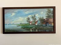 Framed landscape canvas print showing three houses near a waterbody with trees and a windmill in the background under blue sky.