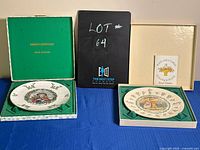 Two Royal Doulton collectible plates with original packaging placed on a blue cloth background, showing the plates inside their boxes and an information leaflet.