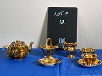 A rich brown and gold mottled small teapot with lid, a handled basket on a square base, a creamer, a cup and a saucer arranged on blue cloth with black sign showing lot number 12.