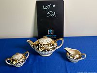 Gold and white Sadler teapot, creamer, and sugar bowl against blue background, black board behind.