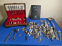 Wide shot showing the wooden cutlery case with red fabric interior holding some forks, spoons, and knives; the rest of the cutlery is arranged on a blue cloth surface with visible tarnish.