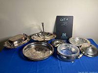 Seven silver-plated items including trays, covered dishes, and a two-tiered stand displayed on a blue cloth with visible tarnish.