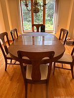 Front view of the dining table with 6 chairs arranged around it near a window showing the polished wood surface and oval shape.