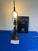 Full view of the vintage metal lamp with bulb lit, showing the base, harp, and finial with a note in front.