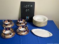 Full view of all items including 4 blue and orange consomme bowls with saucers, stack of 12 white bowls with gold trim, and white serving tray on blue table.