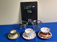 Photo of three vintage teacups with matching saucers and two glass candy dishes on blue cloth background.