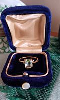 Ring in open blue velvet box displaying emerald-cut light green stone and gold setting