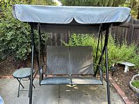 Full view of the 2-person black metal mesh swing with attached gray fabric canopy and small round side table.