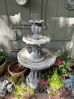 Full view of two-tier gray resin garden fountain surrounded by plants and placed beside a wall.