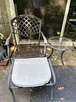 One aluminum outdoor chair with lattice back, polyester cushion, next to round glass top table with aluminum frame.