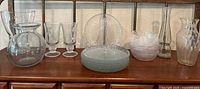 Lot arranged on wooden shelf showing various clear glassware and plastic tableware items including glasses, bowls, plates, vases, and pitcher.