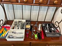 Wide view of assorted kitchen utensils on wooden shelf with several white and brown trays containing various flatware, peelers, scissors, measuring spoons, clips, graters, and other items.