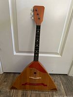 Full front view of the balalaika showing triangular wooden body, fretted neck, headstock, strings, and red painted decoration and text.