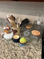Photo showing kitchen utensils in ceramic grapevine holder, glass canisters with wooden lids, ceramic chicken dish, and two cupcake-shaped containers.