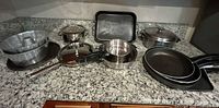 Multiple cookware pieces on granite countertop, showing pots, pans, roasting tray, metal tubs, and lids.