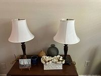 Full lot overview showing 2 metal base lamps with white shades, 3 vases in beige, gray and blue, rectangular ceramic vase with white artificial flowers, and a small framed painting on wooden table.