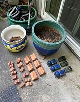 Photo showing three ceramic pots including a plain green one filled with garden hose and a lawn sprinkler, a smaller sunflower painted pot, and a medium blue-green pot with soil inside. Various terracotta pot feet and glazed pot feet are arranged in front.