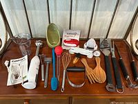 Kitchen utensils and gadgets laid out showing variety of items from immersion blender, kitchen bowl, measuring spoons, whisks, tongs, and peelers.