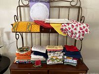 Large lot of folded kitchen linens on metal rack and wooden surface, including dish towels, napkins, and placemats with various colors and patterns