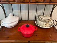 Three items displayed on wooden surface: red Dutch oven centrally positioned, white covered casserole dish to left, covered stoneware pot to right