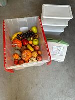 Assorted artificial fruits in a clear plastic storage bin with red handles, including pumpkins, apples, pears, bananas, grapes, and other vegetables