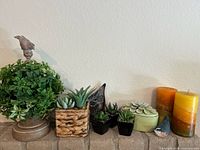Photo showing an artificial green foliage plant on pedestal, four small artificial succulents in woven and black ceramic pots, large ceramic bird sculpture, three-layer pillar candles, and small resin bird figurines.