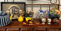 Wide view showing the ceramic teapot with blue and yellow knitted cozy, yellow covered jar, painted wood spoons, small ceramic creamer, and egg cups on display on wooden surface with floral decoration behind