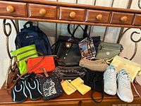 Lot arranged on a table below a wooden console table. Visible are various bags including a navy Affinity backpack, black leather purse, orange clutch, yellow and green wallets, black sandals, and blue sneakers.