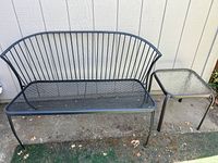 Black metal 2-seater outdoor patio bench with mesh seat and vertical slat backrest positioned beside the side table.
