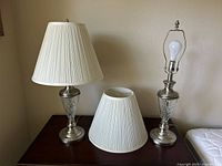 Two silver metal and glass table lamps with cream pleated shades on a brown surface. One lamp has a conical shade, the other shade is removed, revealing light bulb and finial.