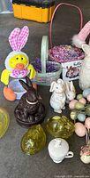 Collection of spring and Easter decor items including plush chick figure with bunny ears, ceramic bunny figurines, glass egg containers, decorative eggs, and baskets with shredded paper filler.