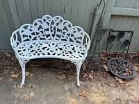Front view of white cast iron garden bench showing floral rose and leaf design with three legs visible, placed near a fence outdoors.