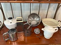Kitchen utensils including salad spinner, metal grater, flour sifter, colanders, sieve on wooden surface