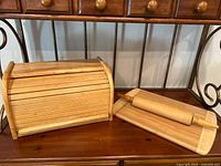 Wooden roll-top bread box closed, medium wooden cutting board, and wooden rolling pin displayed on a wood surface.