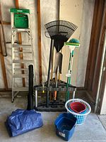 Full view of yard tools, ladder, buckets, tarps, with plastic yard tool rack holding rake, shovel, and broom.