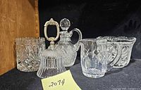 Five pieces of crystal cut glass on display including a bell, decanter, creamer, and sugar bowls, arranged on a black surface with a sticky note showing lot number.