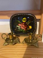 Photo showing two brass ornate candlestick holders placed side by side in front of a floral painted tray with black background and colorful flowers.