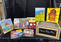 Photo shows a collection of 9 children's books in a woven wooden basket with a chalkboard sign on front reading 'Please Take One'. Titles visible include 'The Not Very Merry Pout-Pout Fish', 'Love Monster', 'Leo the Late Bloomer', 'My Very Favorite Book in the Whole Wide World', and several dragon-themed books.