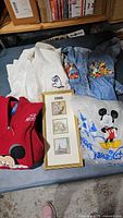 Photo showing the full lot laid out on a surface, includes Winnie the Pooh denim dress, Eeyore white shirt, red Mickey vest, gray Disney sweatshirt, and framed art with three illustrations.