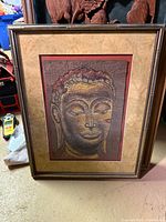 Framed batik painting of Buddha face on textured gold and red-brown background.