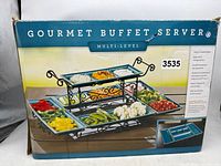 Box front image showing the gourmet buffet server with stoneware trays and metal stand containing various food items.