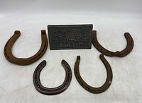 Front view of four metal horseshoes and a rectangular metal bacon press with pig design and lettering.