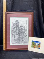 Framed black-and-white sketch print of Rouen, France with brown wood frame and dark red mat beside smaller watercolor painting of Arc de Triomphe