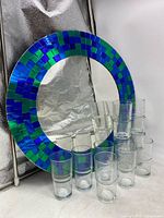 12 clear glass stacking juice glasses arranged in front of round mosaic mirror with blue and green tiles.