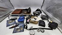 Wide view showing all items in the lot including vintage cameras, camera cases, binoculars, camera strap, small film canisters, Metal Earth model kits, and brochures.